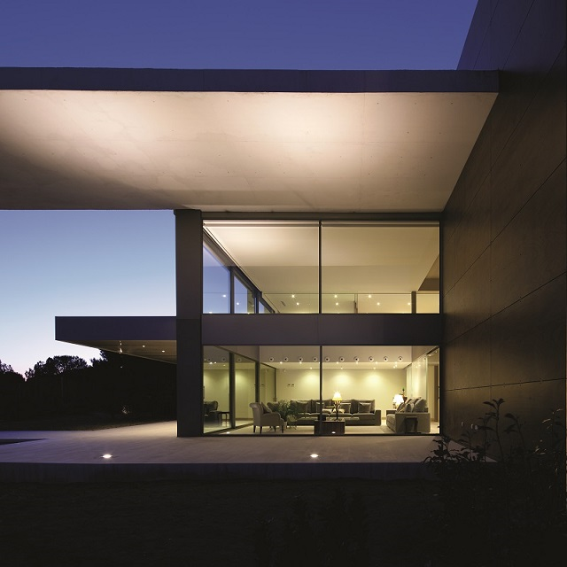 a square modern building is shown at sunset. There is large glass walls showing a view  into the property.