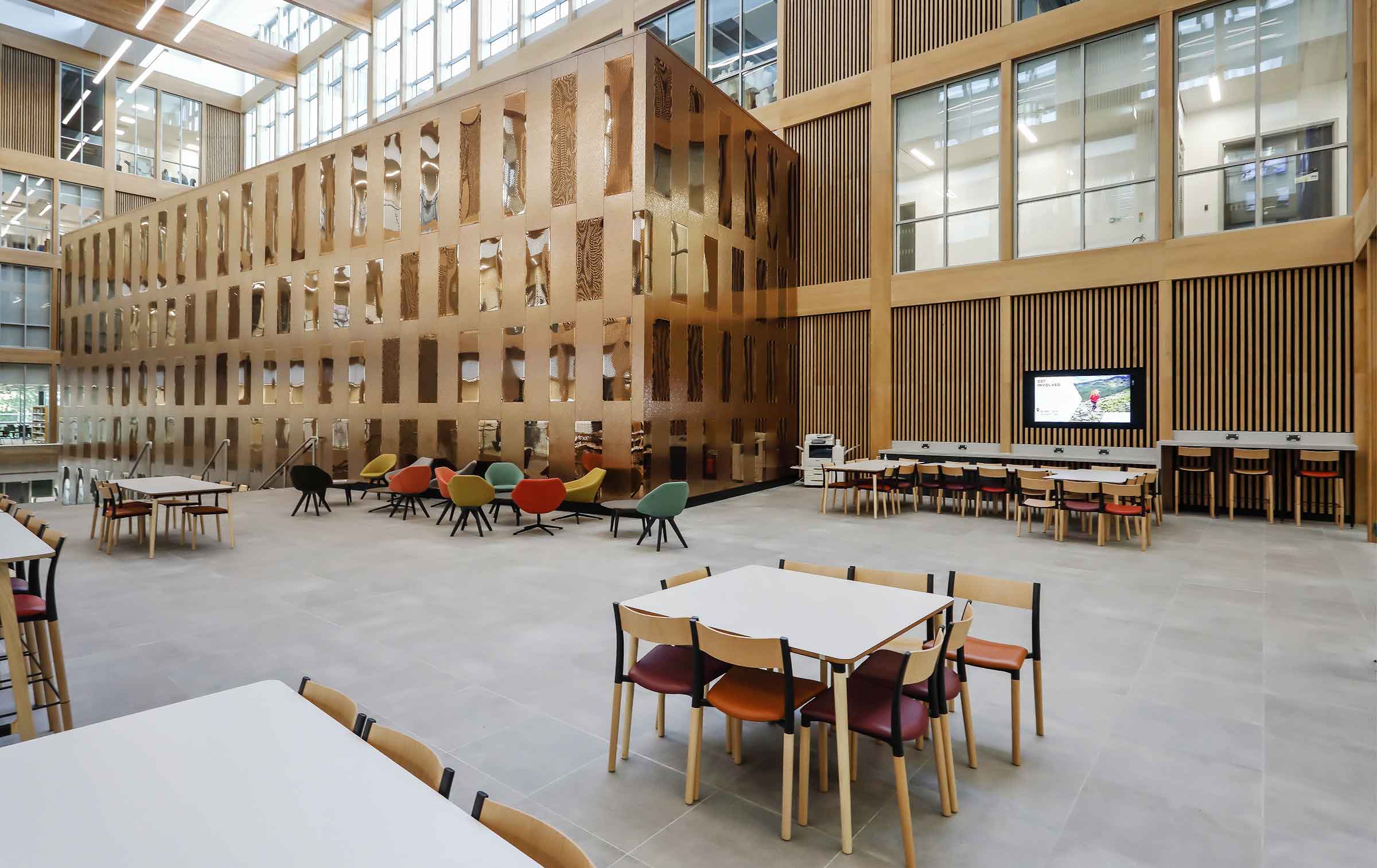 The inside of a modern educational building. There is a gold-colour, square building to the rear of the shot and a glazed wall. To the front, there are study spaces with chairs around a table.