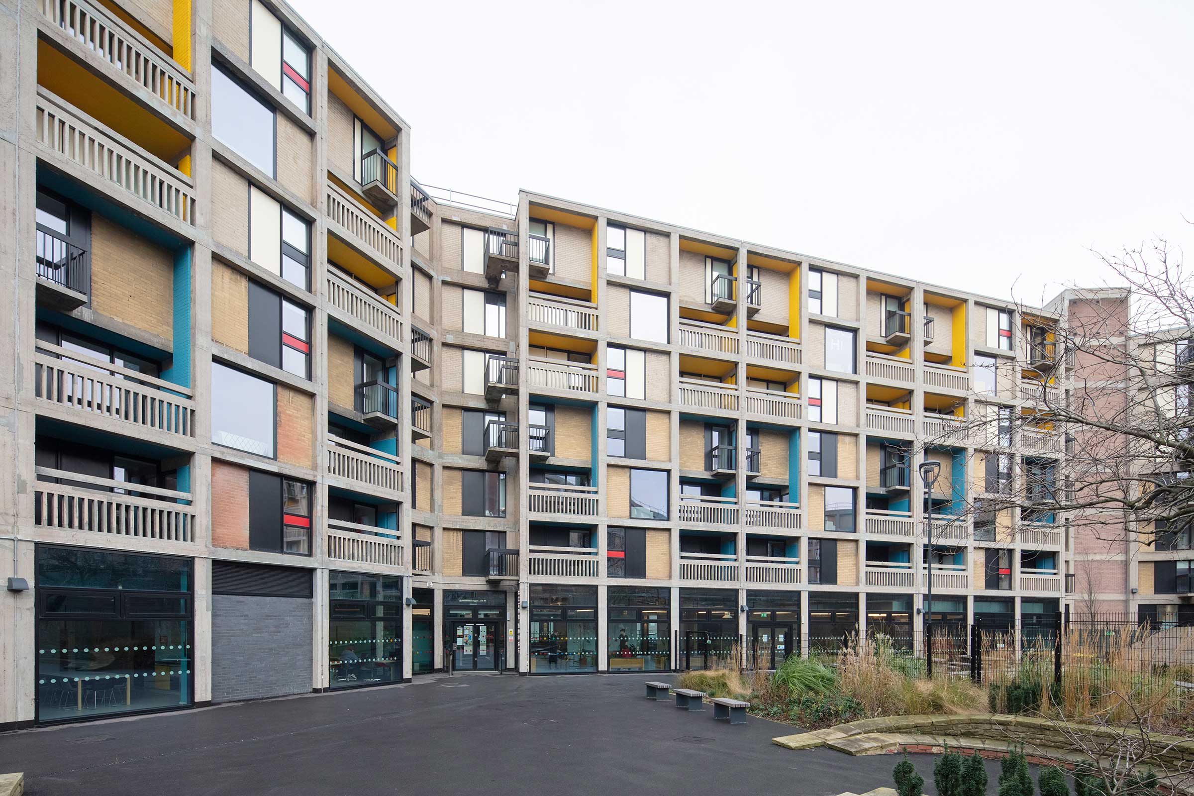 the external of a brutalist apartment block that has been refurbished with new aluminium windows. There are blocks of blue, red and yellow infill panes to the sides of the building.