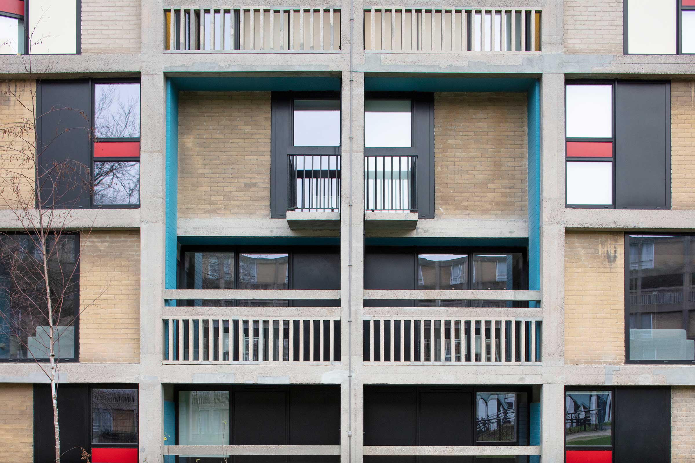 a front facing image of a brutalist, concrete building. There are aluminium windows and coloured panels interspersed between brick.