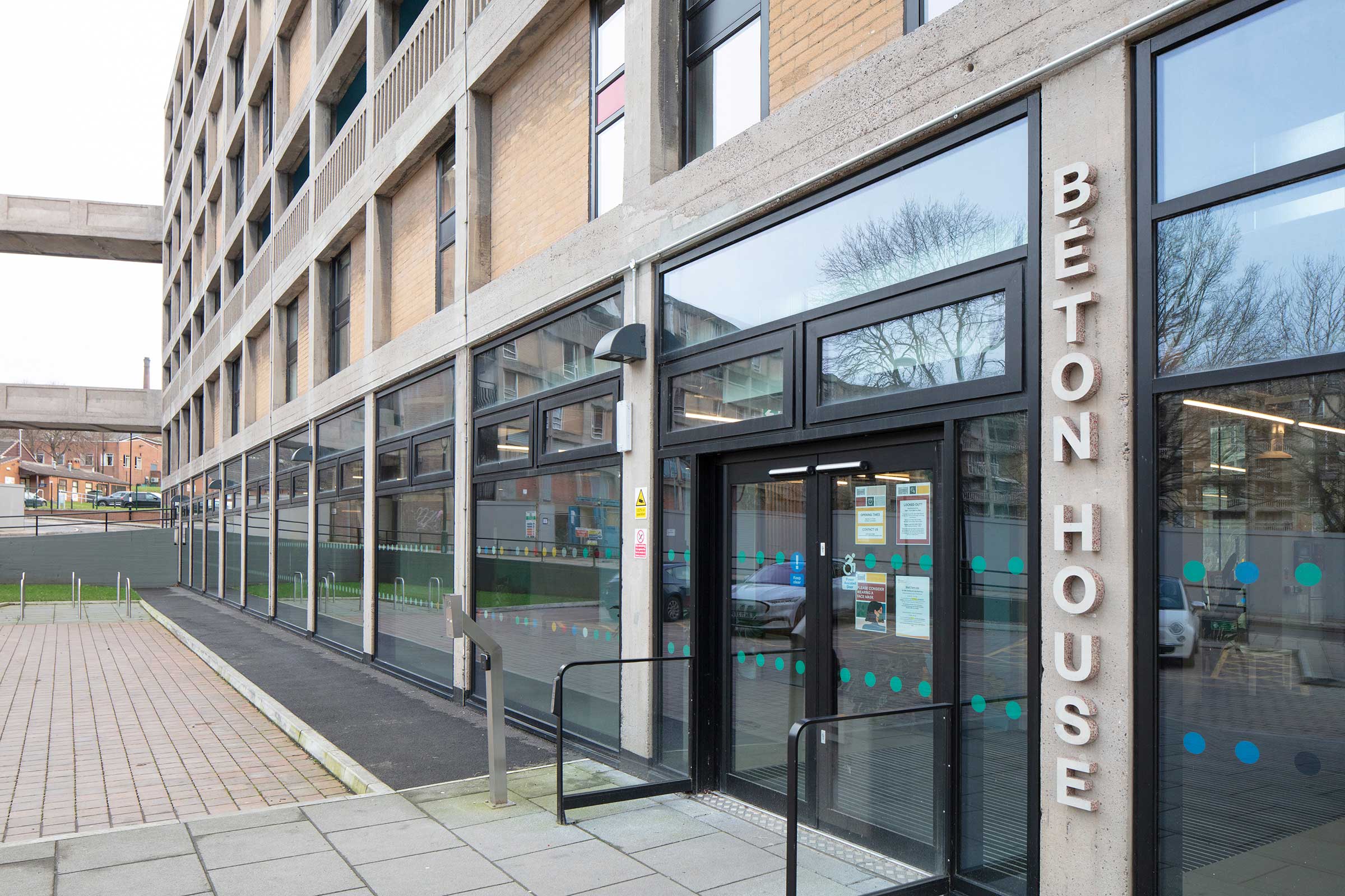 An image of an apartment block with concrete and brick walls and aluminium windows, doors and facades, There is a sign next to the entrance door which reads "Beton House",